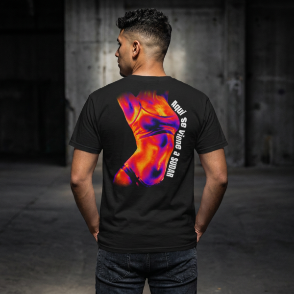 Hombre de espaldas en un entorno industrial oscuro vistiendo una camiseta rave negra oversize. La prenda tiene un estampado trasero de mapa térmico de un torso femenino y el texto vertical "Aquí se viene a sudar" en blanco.